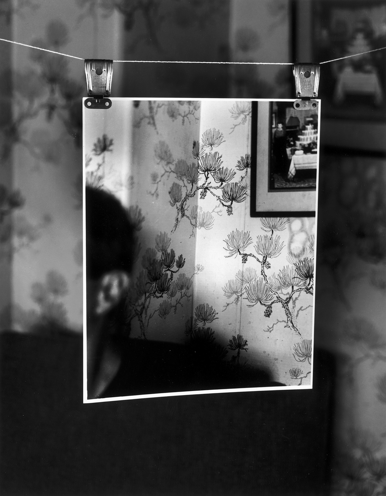 A photograph of the room in the background, with distinctive wallpaper and a framed photograph on the wall, is suspended from a string by two clips in the foreground. The photograph in the foreground has part of the side of a person’s face that is not visible in the background.